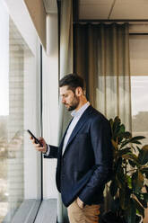 Side view of businessman using smart phone while standing near window in coworking office - MASF38061