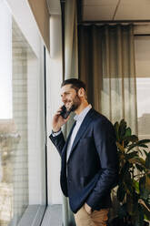 Smiling businessman talking on mobile phone while standing near window in coworking office - MASF38062