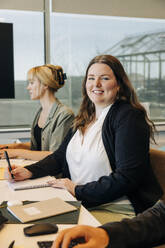 Porträt einer lächelnden Geschäftsfrau, die mit Kollegen am Schreibtisch in einem Coworking-Büro sitzt - MASF38077