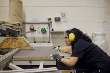 Side view of female carpenter wearing protective eyewear and ear protectors while working in factory - MASF38227
