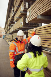 Mature female worker discussing with colleague while standing by stack of planks - MASF38262