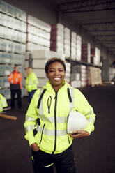 Portrait of smiling female worker in reflective clothing holding hardhat standing in factory - MASF38273