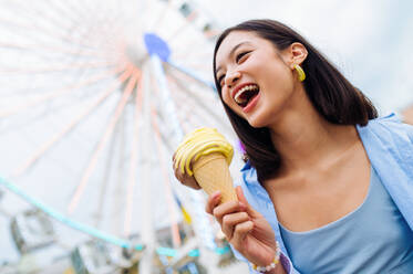 Beautiful young asian woman eating ice cream at amusement park - Cheerful chinese female portrait during summertime vacation- Leisure, people and lifestyle concepts - DMDF03442