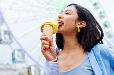 Beautiful young asian woman eating ice cream at amusement park - Cheerful chinese female portrait during summertime vacation- Leisure, people and lifestyle concepts - DMDF03444