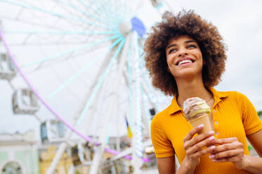 Beautiful young black woman eating ice cream at amusement park - Cheerful african-american female portrait during summertime vacation- Leisure, people and lifestyle concepts - DMDF03445