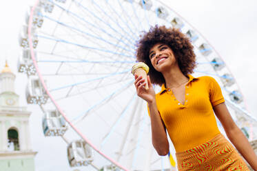 Beautiful young black woman eating ice cream at amusement park - Cheerful african-american female portrait during summertime vacation- Leisure, people and lifestyle concepts - DMDF03447