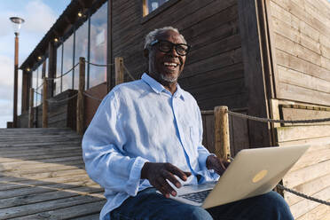 Happy engineer sitting with laptop in front of cafe - ASGF04467