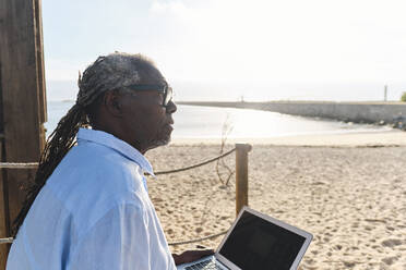 Nachdenklicher Ingenieur sitzt mit Laptop am Strand an einem sonnigen Tag - ASGF04468