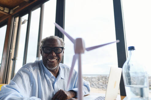 Happy senior engineer with wind turbine model sitting in cafe - ASGF04473