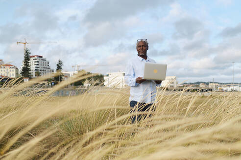 Ingenieur steht mit Laptop inmitten von langem Gras auf einem Feld - ASGF04508