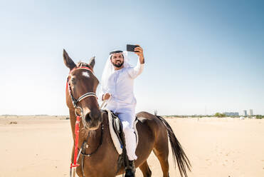 Middle eastern handsome man with typical emirates dress riding a arabic horse in the Dubai desert - DMDF03618