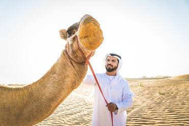 Handsome middle eastern man with kandura and gatra riding on a camel in the desert - DMDF03624