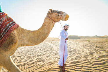 Handsome middle eastern man with kandura and gatra riding on a camel in the desert - DMDF03625