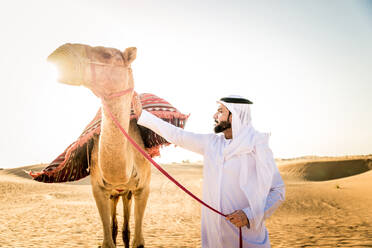 Handsome middle eastern man with kandura and gatra riding on a camel in the desert - DMDF03626