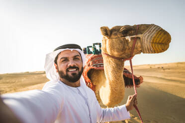 Handsome middle eastern man with kandura and gatra riding on a camel in the desert - DMDF03628