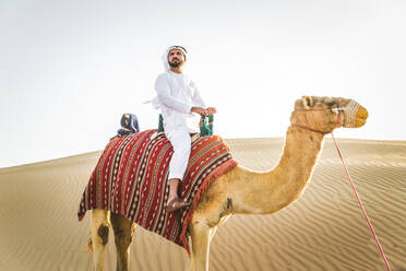 Handsome middle eastern man with kandura and gatra riding on a camel in the desert - DMDF03629