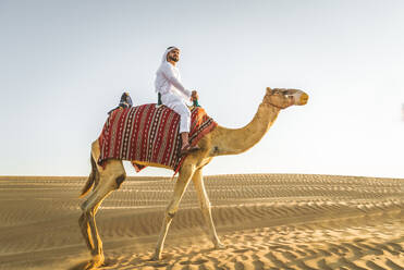 Handsome middle eastern man with kandura and gatra riding on a camel in the desert - DMDF03630