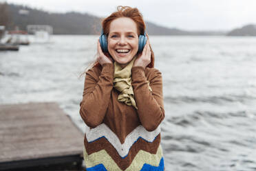 Fröhliche Frau hört Musik über drahtlose Kopfhörer am See - KNSF09744