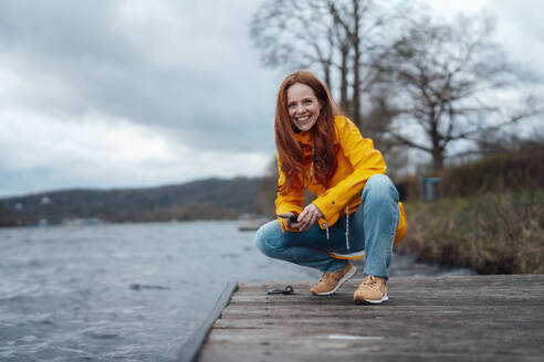 Smiling redhead woman squatting on pier by lake - KNSF09750