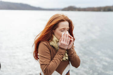 Redhead woman blowing nose in front of lake - KNSF09751