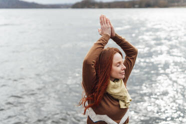 Rothaarige Frau beim Yoga mit verschränkten Händen am See - KNSF09756