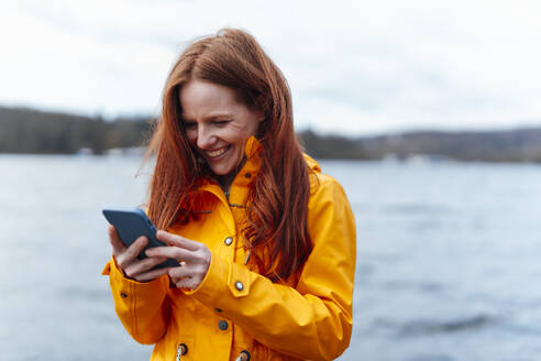 Glückliche rothaarige Frau mit Smartphone vor einem See - KNSF09767