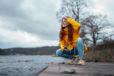 Smiling woman squatting on pier by lake - KNSF09775