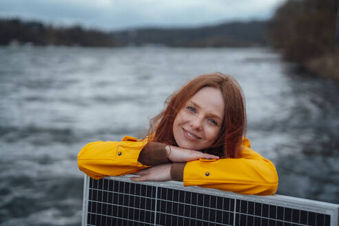Smiling redhead woman leaning on solar panel in front of lake - KNSF09780