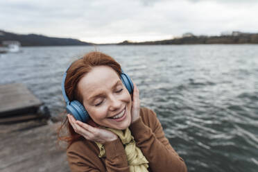 Lächelnde Frau hört Musik über drahtlose Kopfhörer am See - KNSF09785