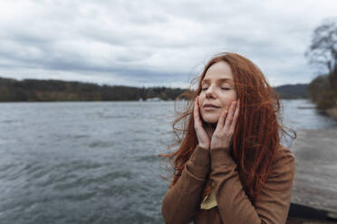 Woman touching face with eyes closed in front of lake - KNSF09799