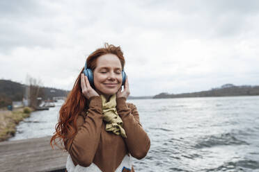 Smiling woman listening to music with eyes closed by lake - KNSF09800