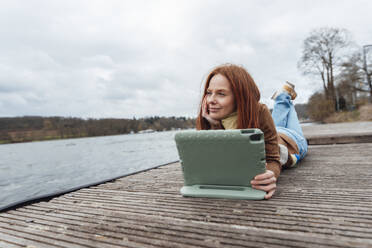 Kontemplative Frau mit Tablet-PC auf dem Pier am See liegend - KNSF09808