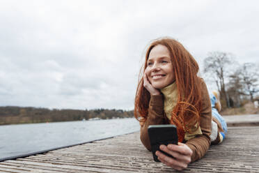 Lächelnde Frau mit Mobiltelefon auf dem Pier am See liegend - KNSF09809