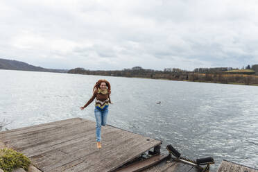 Carefree woman jumping on pier by lake under cloudy sky - KNSF09810