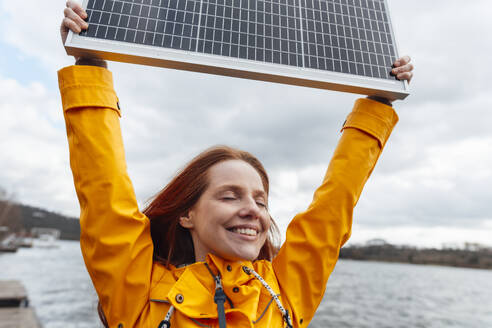 Lächelnde Frau mit geschlossenen Augen, die ein Solarpanel hält - KNSF09817