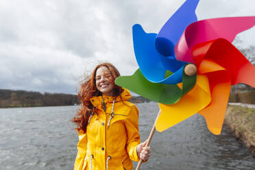 Smiling woman with pinwheel toy in front of lake - KNSF09822