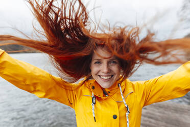 Happy redhead woman with tousled hair by lake - KNSF09826