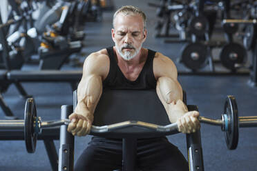 Älterer Mann, der seine Arme mit einer Langhantel im Fitnessstudio stärkt - KPEF00215