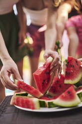 Hands of friends picking up slices of watermelon at table - ANNF00418