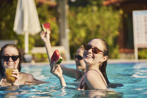 Glückliche Frau hält ein Stück Wassermelone in der Hand und genießt es mit Freunden im Pool - ANNF00448