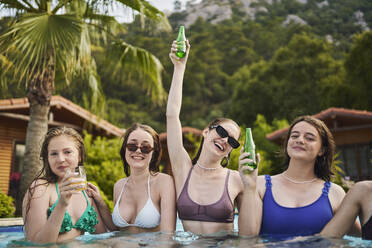 Unbeschwerte Frau hält Bierflasche mit Freunden, die sich im Pool vergnügen - ANNF00469