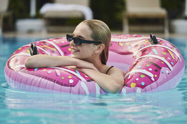 Lächelndes jugendliches Mädchen mit aufblasbarem Schwimmring, das sich im Pool entspannt - ANNF00475