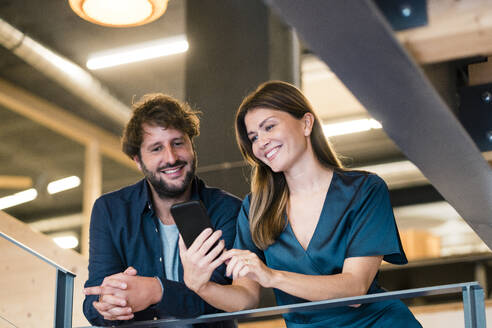 Lächelnde Geschäftsfrau zeigt einem Geschäftsmann im Büro sein Smartphone - JOSEF20563