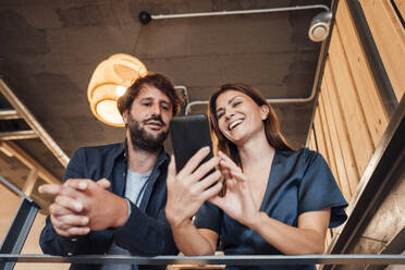 Glückliche Geschäftsfrau, die einem Geschäftsmann im Büro ihr Mobiltelefon zeigt - JOSEF20591