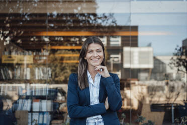 Smiling businesswoman wearing blazer standing in front of glass - JOSEF20595