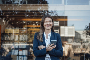 Smiling businesswoman holding smart phone in front of glass - JOSEF20597