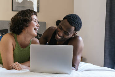 Lächelndes Paar auf dem Bett liegend mit Laptop zu Hause - PBTF00165