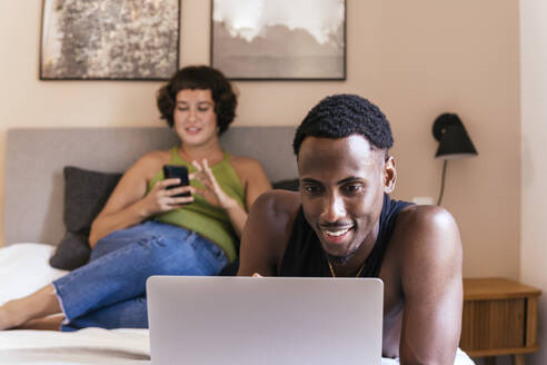Lächelndes Paar mit Laptop und Smartphone auf dem Bett zu Hause - PBTF00167
