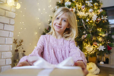 Lächelndes blondes Mädchen mit Weihnachtsgeschenk zu Hause - NJAF00576