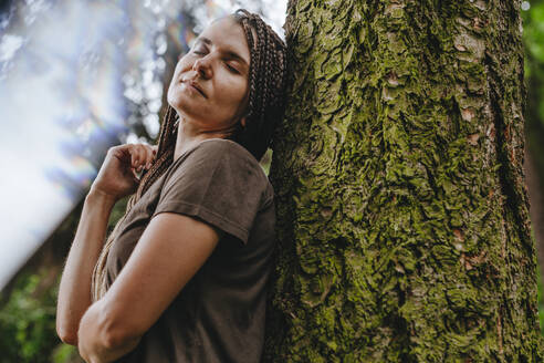 Frau mit geschlossenen Augen lehnt an einem Baum im Wald - YTF01116